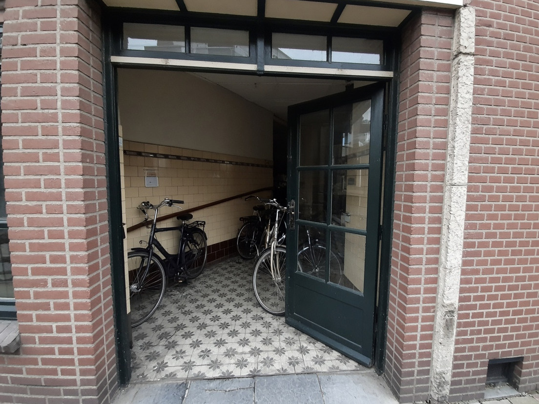 Portiekentree met tegels, fietsen en een glazen deur op een kier